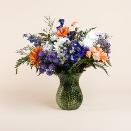 Mixed bouquet of orange, white, blue, and purple flowers in a green glass vase