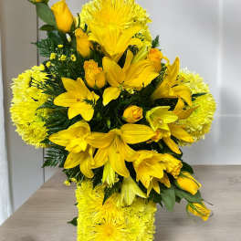 Yellow cross-shaped sympathy arrangement with lilies, roses, and chrysanthemums on a wooden stand