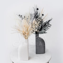 Two dried floral arrangements in white and black vases on a round table.