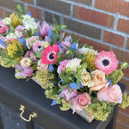 Long wooden box arrangement with pink gerbera daisies, roses, dahlias, blue hyacinths, and yellow protea.