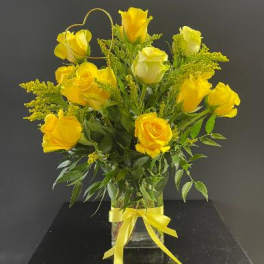 Tall arrangement of yellow roses in a clear glass vase with a yellow bow and heart-shaped accent