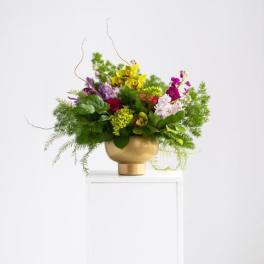 Asymmetrical arrangement of green orchids and mixed bright flowers in a gold pedestal bowl