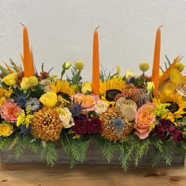 Long floral centerpiece with orange candles and mixed yellow, pink, and burgundy blooms