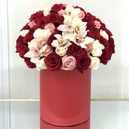 Round arrangement of red, pink, and white roses with orchids in a tall red hatbox-style container