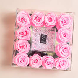 Pink roses arranged in a clear square box around a small center gift box.