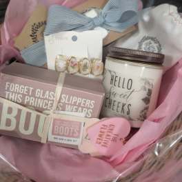 Pastel pink gift basket with candle, earrings, ribbon, and boxed sign set arranged in tissue paper.