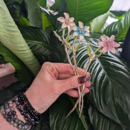 Hand holding three small enamel flower pins against large green leaves