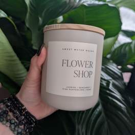 Hand holding a flower shop candle jar in front of large green leaves.
