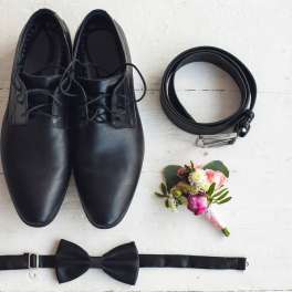 Black dress shoes, belt, bow tie, and a small boutonniere on a white surface