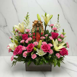 Pink roses and lilies arranged around a religious figurine in a rectangular vase