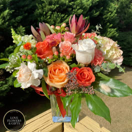 Mixed bouquet of orange, peach, and white flowers in a clear glass vase on a wooden railing