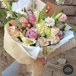 Hand-tied bouquet of pastel roses, lilies, and mixed blooms wrapped in white and burlap-style paper