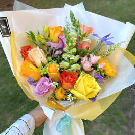 Hand-tied bouquet of yellow, orange, and pink roses with mixed blooms in white wrap.