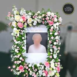 Tall rectangular memorial frame on an easel, bordered with pink and white flowers around a photo area.