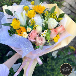 Hand-tied bouquet with yellow sunflowers, roses, white lilies, and pastel roses wrapped in yellow and lilac paper.