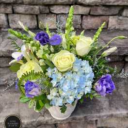 Arrangement of pale yellow roses, blue hydrangeas, and purple blooms in a white vase on stone steps