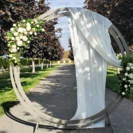 Tall circular metal arch with white flowers and draped white fabric on an outdoor path