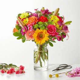 Bright mixed bouquet with sunflower, roses, lilies, and daisies in a clear glass vase on a white surface