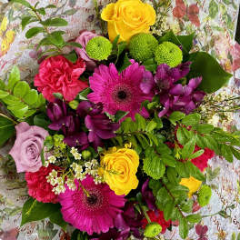 Bright hand-tied bouquet with pink gerbera daisies, yellow roses, green mums, and purple flowers in floral wrap