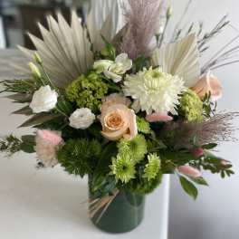 Asymmetrical arrangement of white and peach blooms with green accents in a dark green vase with dried palm fans.