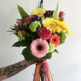Bright hand-tied bouquet of mixed gerbera daisies, roses, tulips, and other flowers with colorful ribbon.
