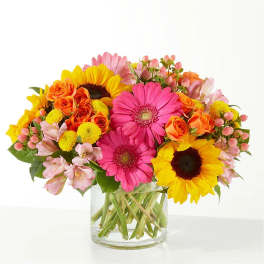 Bright bouquet of pink gerbera daisies, yellow sunflowers, and orange roses in a clear glass vase