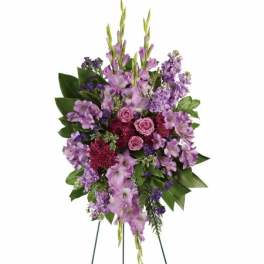 Tall standing spray of purple and pink flowers with roses, gladiolus, and stock on a green leaf backdrop.