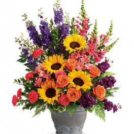 Bright arrangement of sunflowers, orange roses, and mixed pink and purple flowers in a gray pot