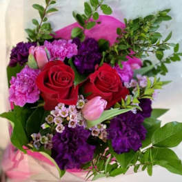 Hand-tied bouquet with red roses, purple carnations, pink tulips, and small white blossoms in pink wrap.