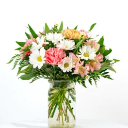 Mixed bouquet of white daisies, pink carnations, and peach blooms in a clear glass jar vase