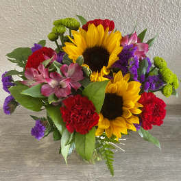 Bouquet with sunflowers, red carnations, pink alstroemeria, and purple filler in a glass vase