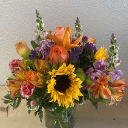 Colorful bouquet with sunflowers, roses, lilies, and purple filler in a glass vase