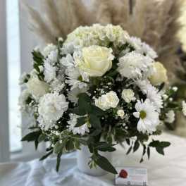 White roses, carnations, and daisies arranged in a white vase with feathery pampas stems in the background.
