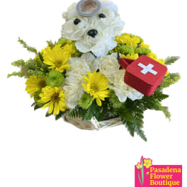 Basket arrangement with white flowers, yellow daisies, and a teddy bear holding a first-aid kit