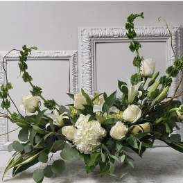 Long low arrangement of white roses, lilies, and hydrangea with tall green accents