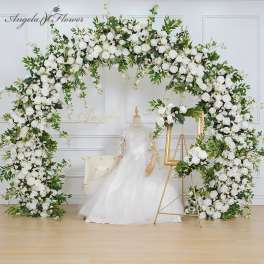 Large white rose wedding arch with matching floral frame beside a mannequin in a bridal gown and chair.