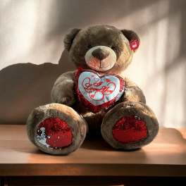 Brown teddy bear with red sequin heart reading "I Love You" on a wooden surface