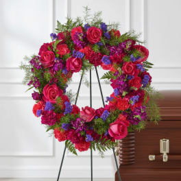 Heart-shaped floral wreath with pink roses and purple flowers on an easel beside a casket