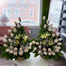 Pair of white rose and lily arrangements with baby's breath in woven baskets on a white table.