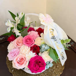 Round basket of pink, red, and white flowers with a congratulatory ribbon