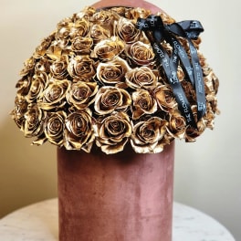 Gold roses arranged in a pink cylindrical box with a black ribbon