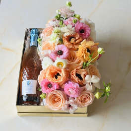 Boxed bouquet with peach, pink, and white flowers beside a bottle of sparkling wine