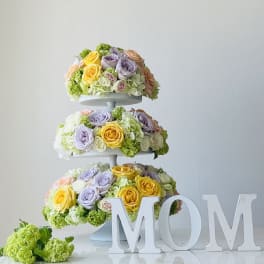 Three-tier floral arrangement with pastel roses and hydrangeas beside large MOM letters