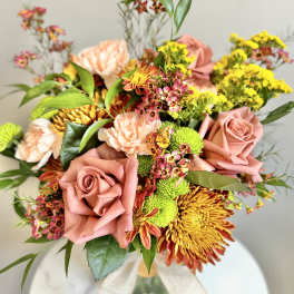 Peach roses, carnations, yellow mums, and green button poms arranged in a clear glass vase with a bow