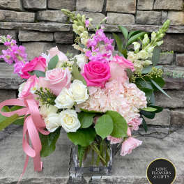 Pink and white roses, hydrangeas, and snapdragons in a clear glass cube vase with a pink ribbon.