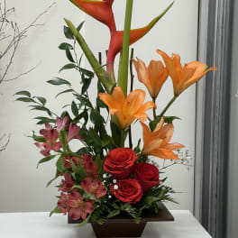 Tropical arrangement with orange heliconia, red roses, and pink alstroemeria in a dark vase