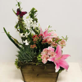 Pink lilies and white orchids in a wooden planter with moss and a butterfly accent
