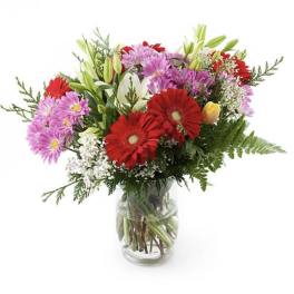 Mixed bouquet of red gerbera daisies, pink flowers, and white lilies in a glass vase