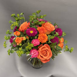 Bright arrangement of orange roses, pink blooms, and purple asters in a clear glass vase