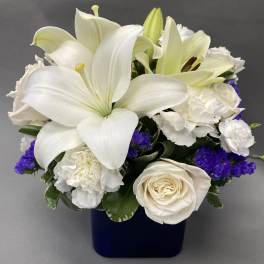 Compact arrangement of white lilies, roses, and carnations with purple accents in a dark blue cube vase